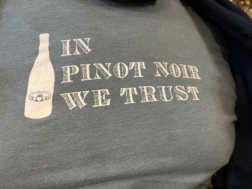 A grey T-shirt, worn by someone whose head is not visible, bearing the text IN PINOT NOIR WE TRUST